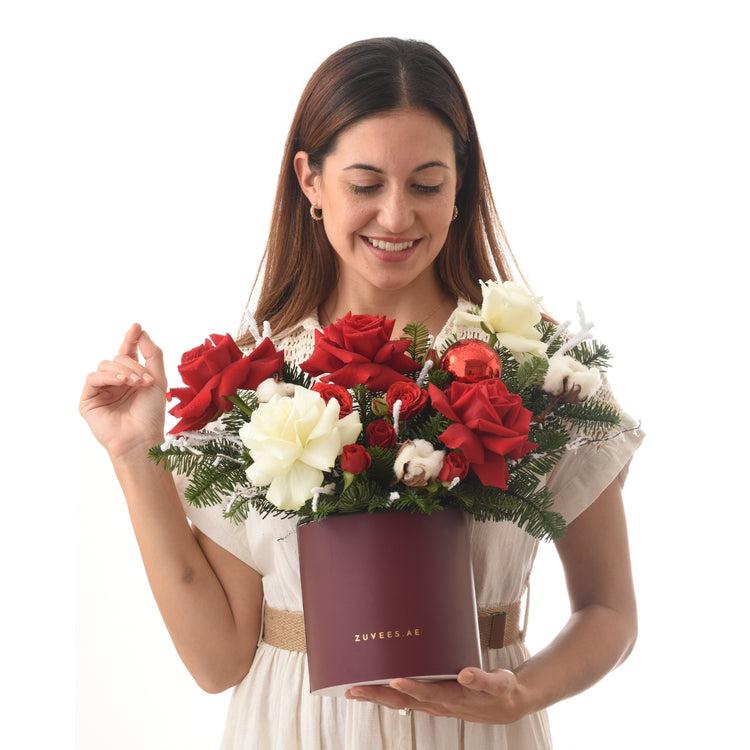 Woman smiles holding burgundy box with red and white roses, pine, cotton, and red ornament.