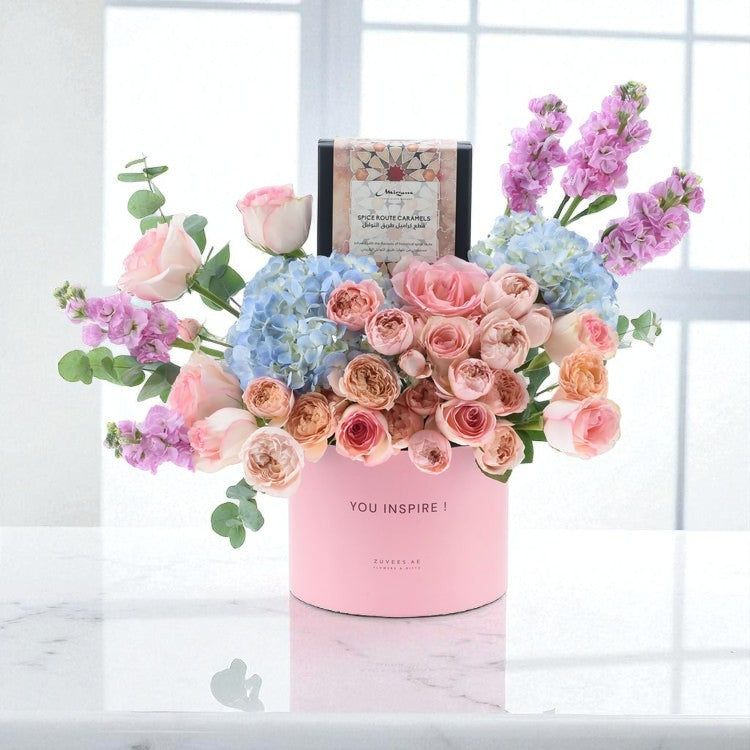 Pastel pink floral arrangement with hydrangeas, roses, and lilacs in branded pink cylinder vase.