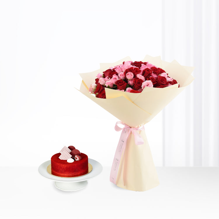 Vibrant red and pink roses bouquet with cream wrap, beside red velvet cake on stand.