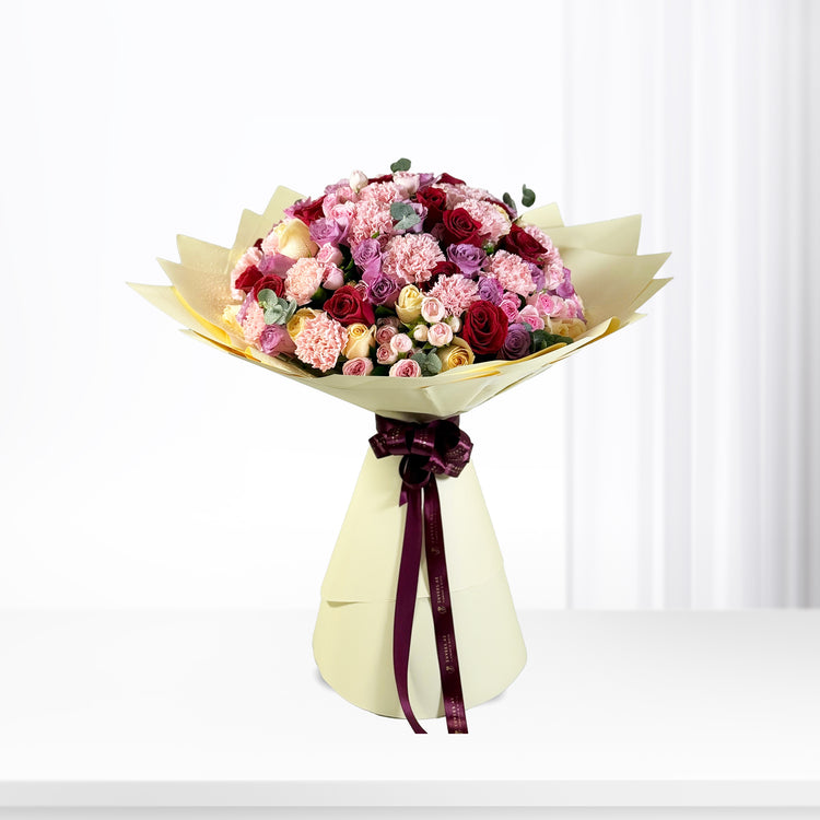 Vibrant pink, red, and yellow roses and carnations wrapped in cream paper with burgundy ribbon.