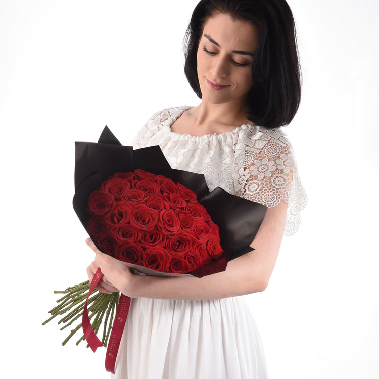 Woman holds vibrant red rose bouquet wrapped in black paper with red ribbon, wearing white lace dress.