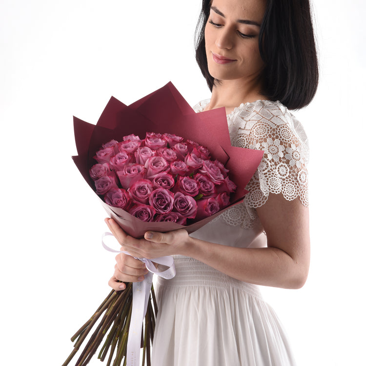 Woman in white lace dress holds burgundy-wrapped bouquet of pink roses.