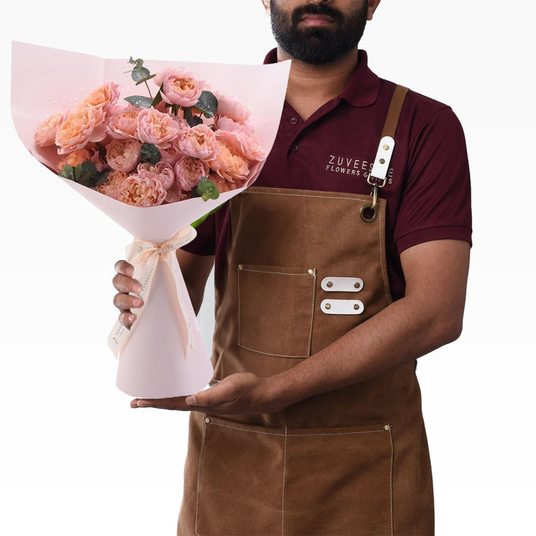 Florist holds lush pink rose bouquet in soft pink wrap, wearing brown apron, maroon polo.