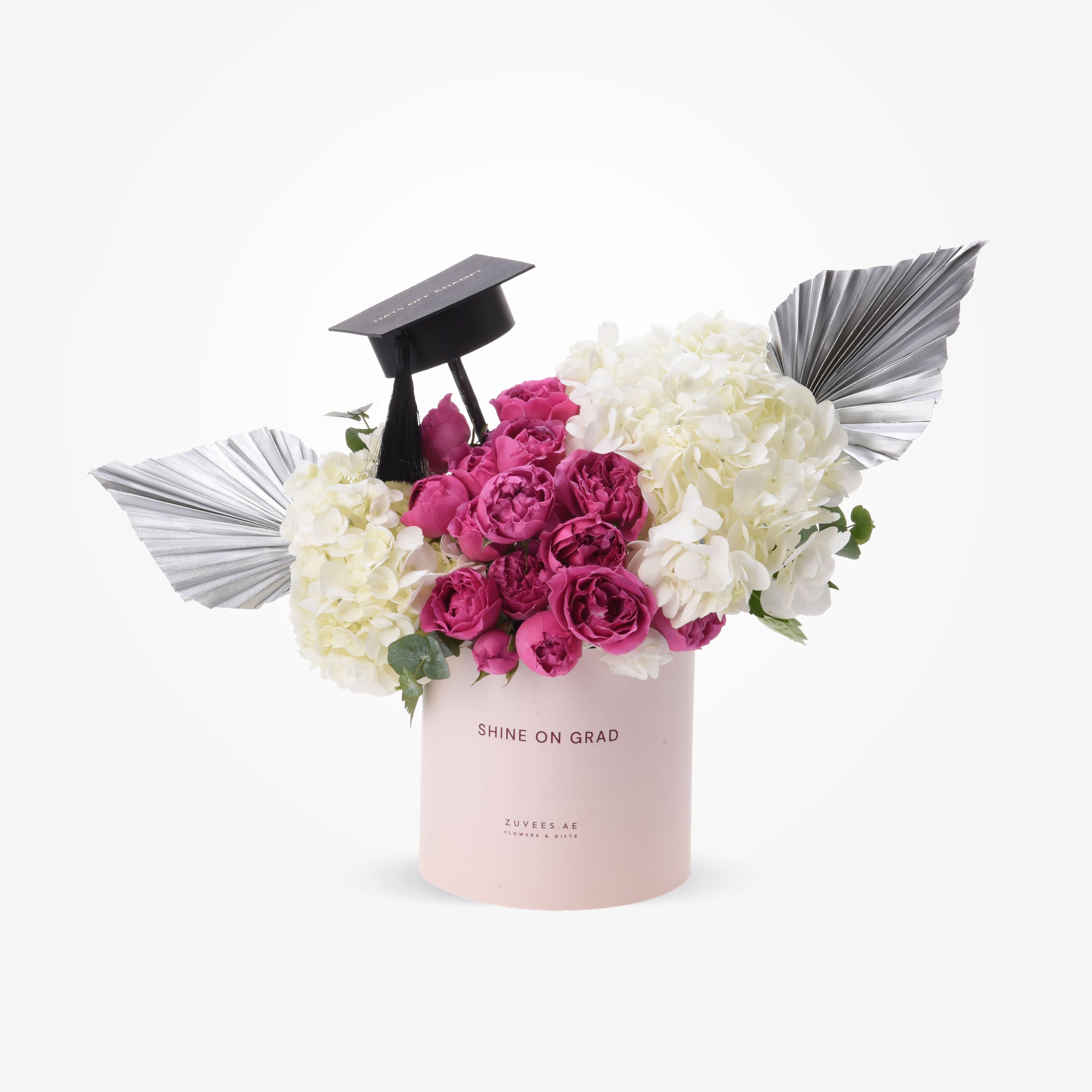 Pink cylinder holds vibrant pink roses and white hydrangeas, accented by silver wings and graduation cap.