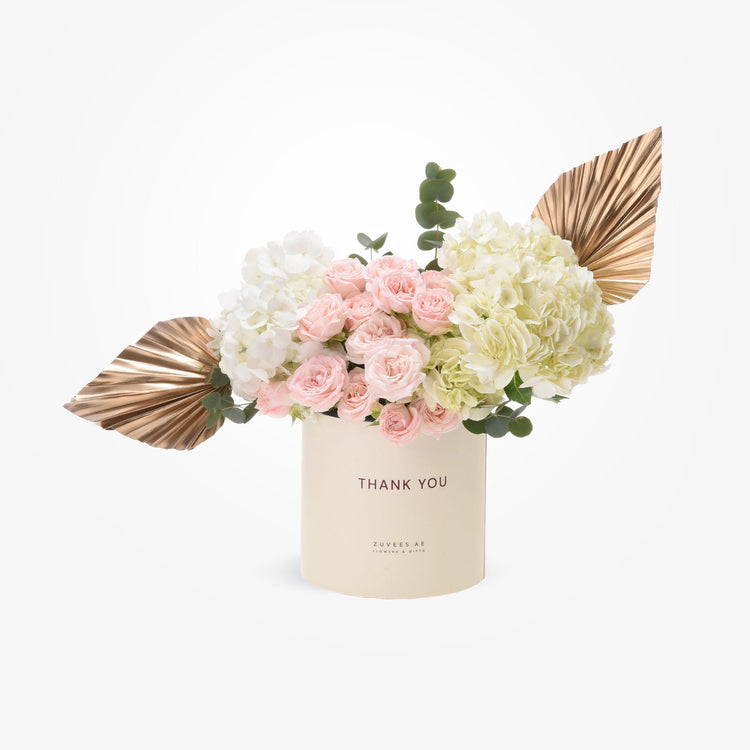 Cream cylinder holds blush roses, white hydrangeas, and gold fan leaves—“Thank You” gift.