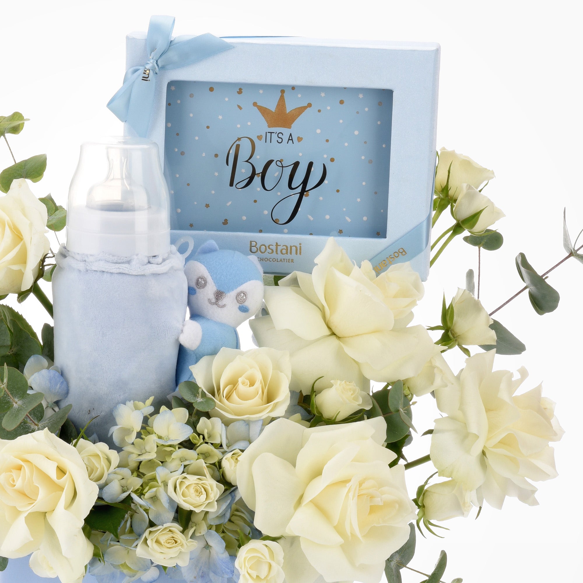 Soft blue baby bottle, plush toy, and chocolate gift box nestled in creamy white roses.