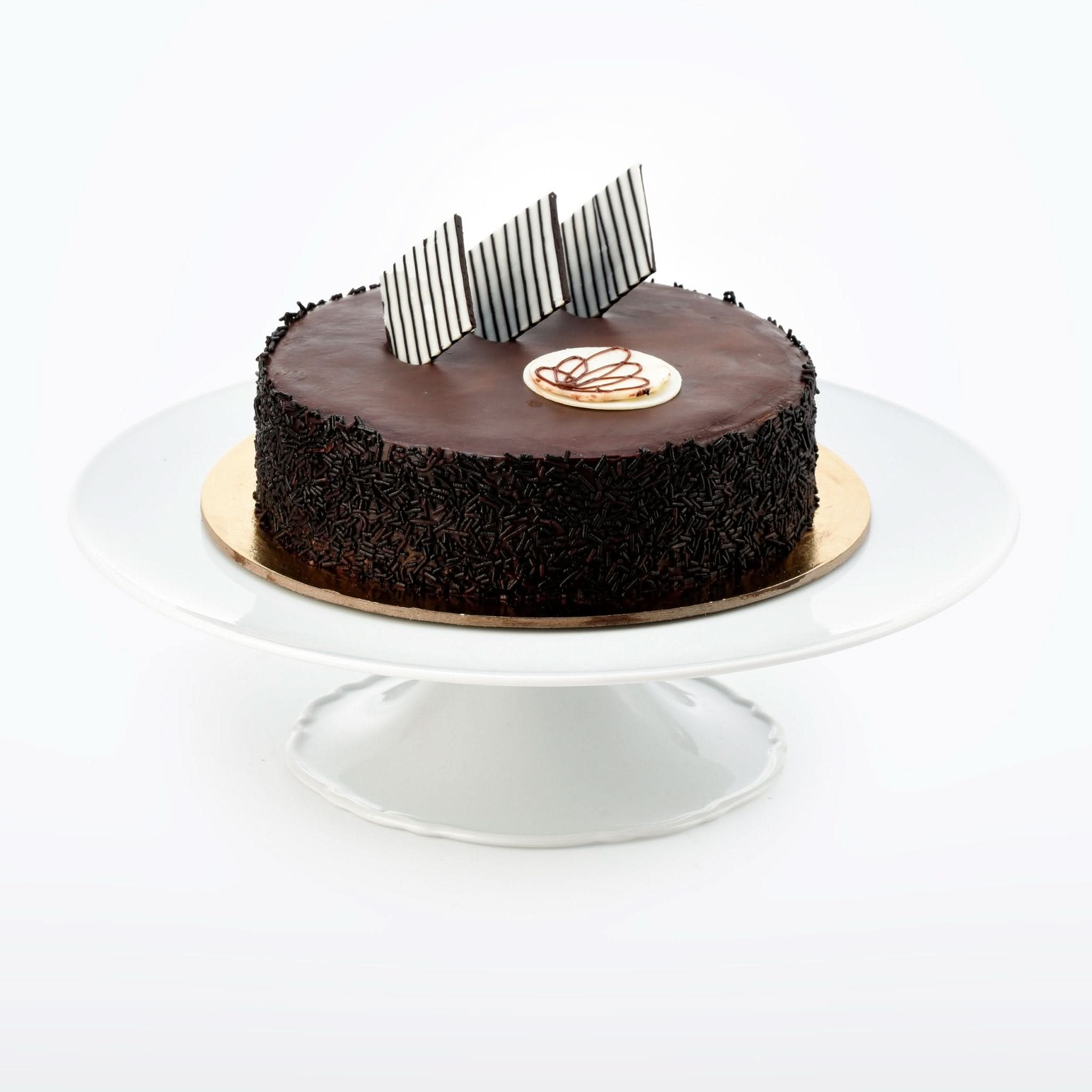Rich chocolate cake adorned with striped chocolate shards and sprinkles on white pedestal.