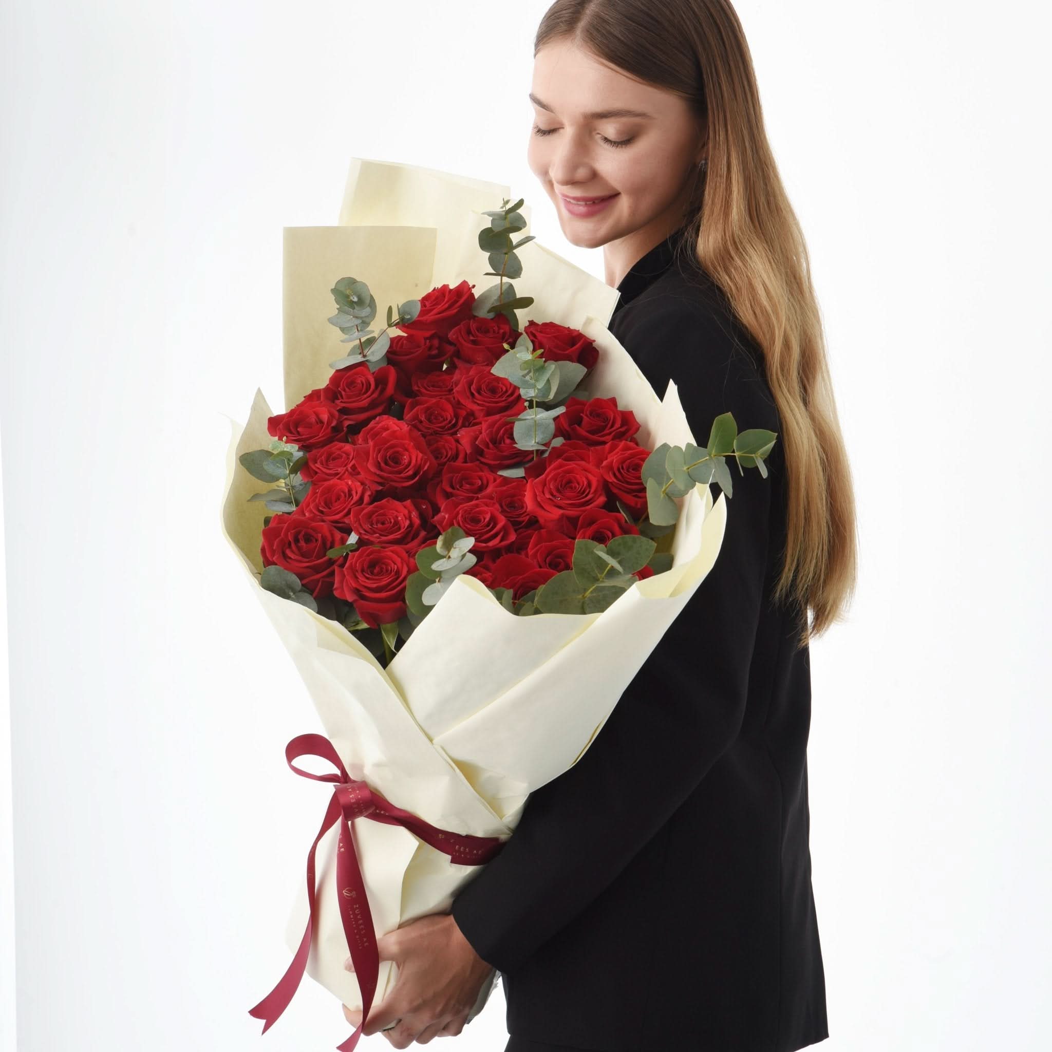 Woman smiles holding vibrant red rose bouquet wrapped in cream paper with burgundy ribbon.