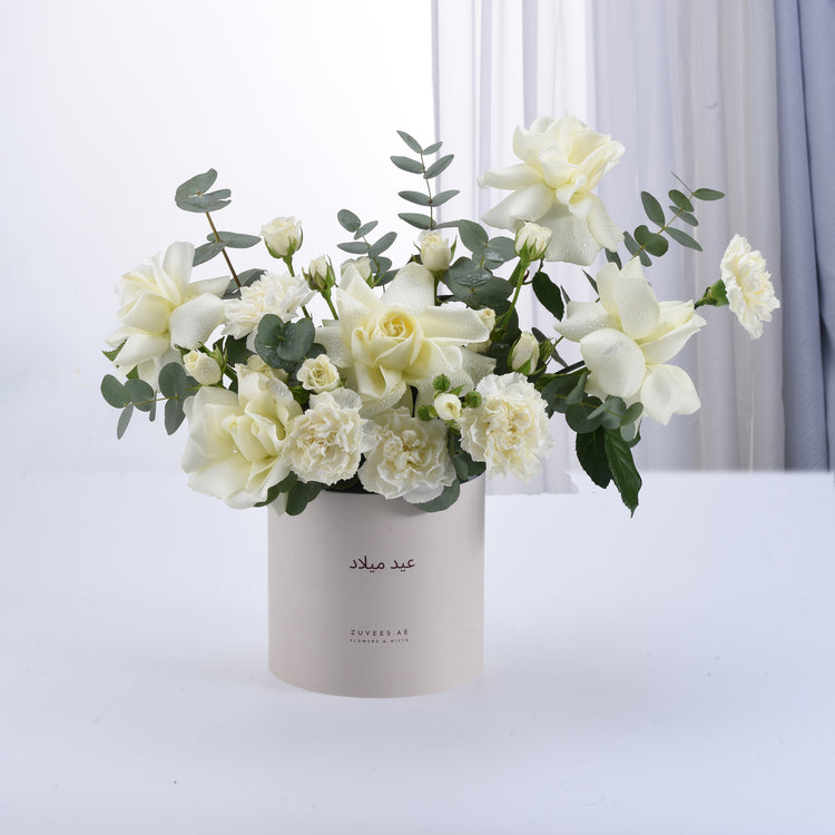 Elegant white rose and carnation bouquet in branded beige cylinder vase.