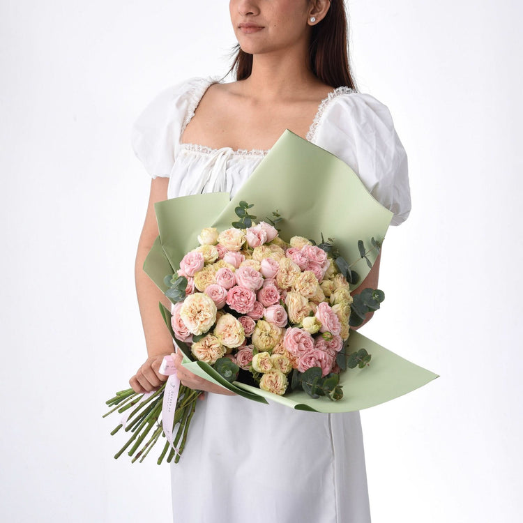 Soft pink and cream roses wrapped in sage green paper, held by a woman in white.