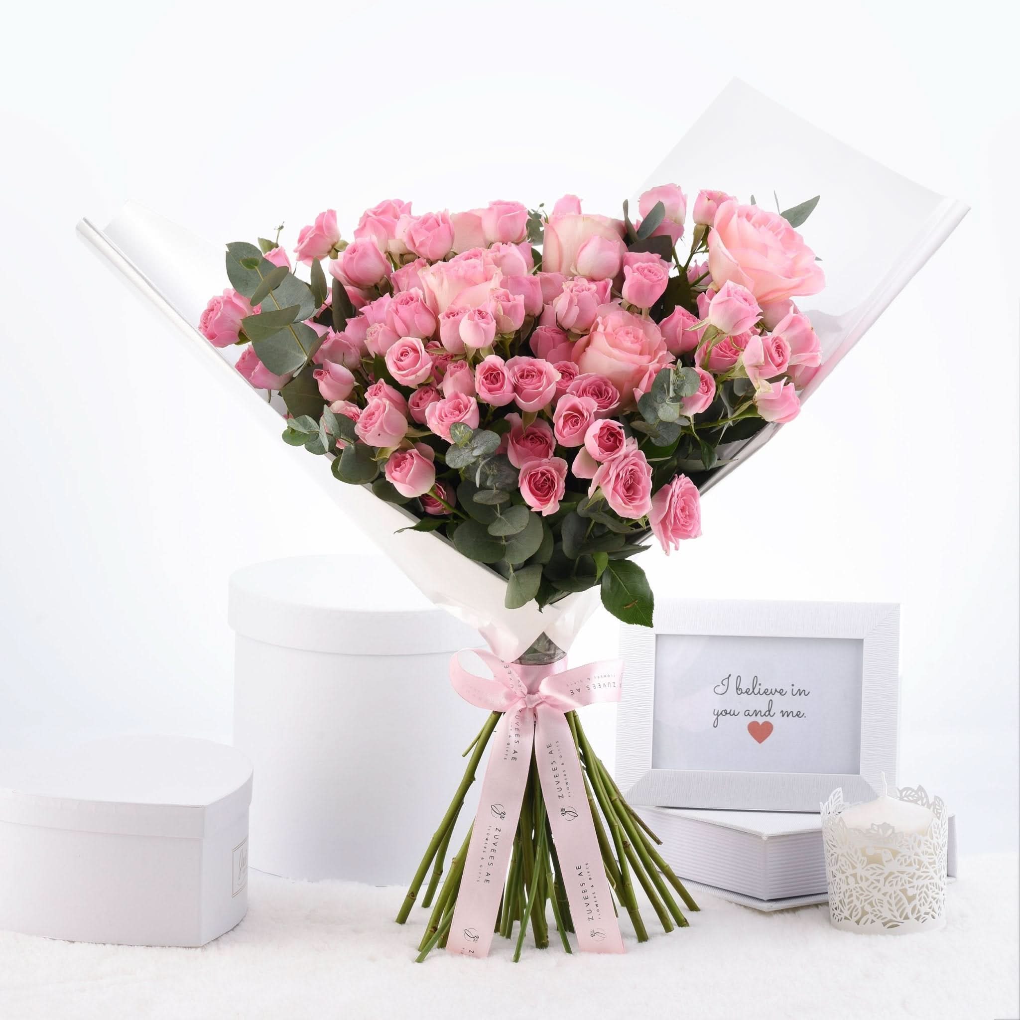 Soft pink roses in white wrap, tied with ribbon, beside framed message and candle.