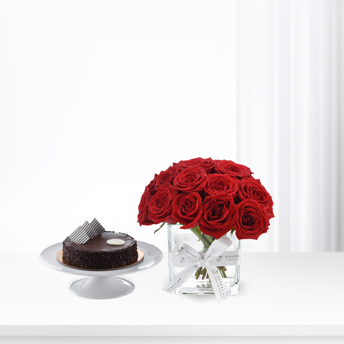 Vibrant red roses in glass vase beside rich chocolate cake on white pedestal.