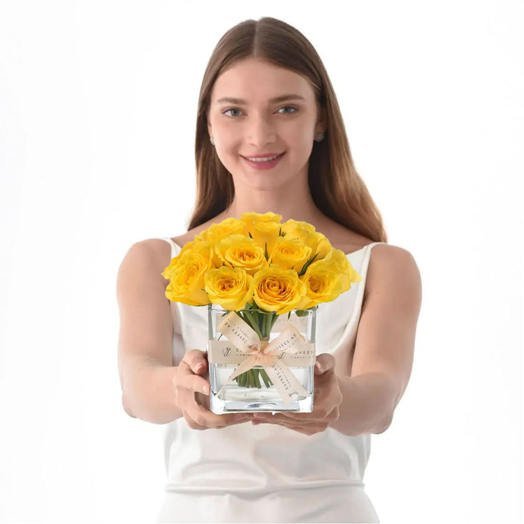 Woman holds clear vase with vibrant yellow roses, tied with cream ribbon.