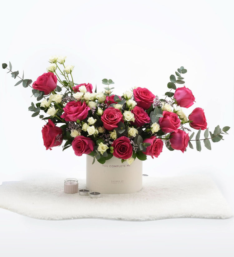 Vibrant pink and white roses in cream cylinder, nestled on fluffy white rug with candles.