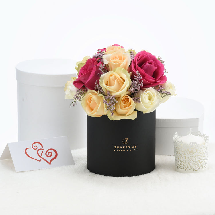 Vibrant pink, peach, and cream roses in black box with gold logo, beside white candle and heart card.