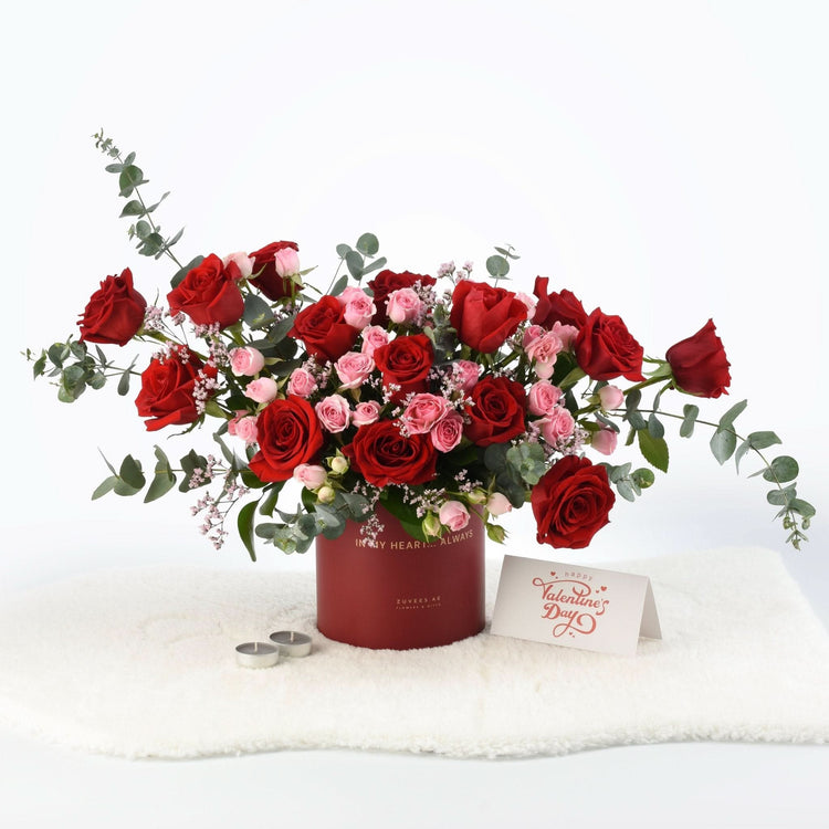 Luxurious red and pink roses in deep red box with Valentine’s card, candles, and eucalyptus on white fur.