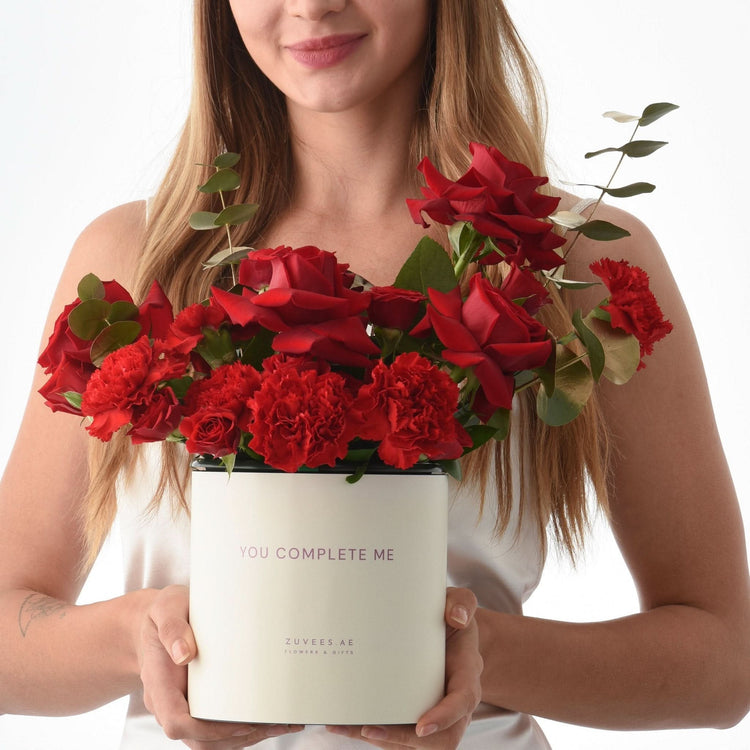 White box holds vibrant red roses and carnations, reads “YOU COMPLETE ME.”