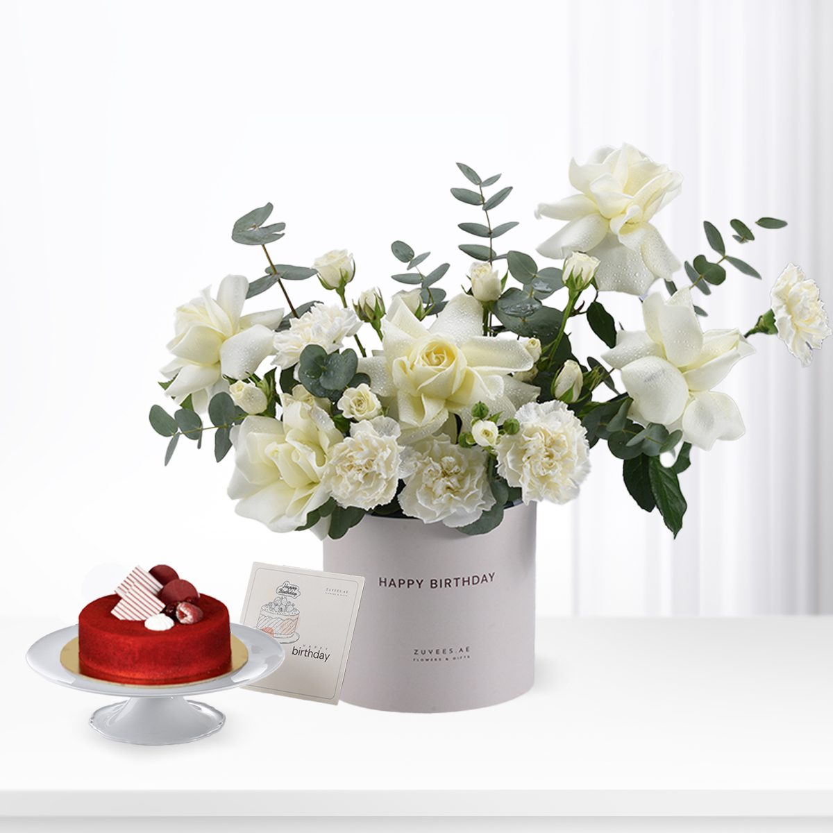 White roses and eucalyptus in gray box beside red velvet cake.