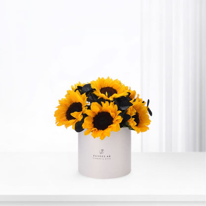 Vibrant sunflowers in matte white cylindrical vase with brand logo.