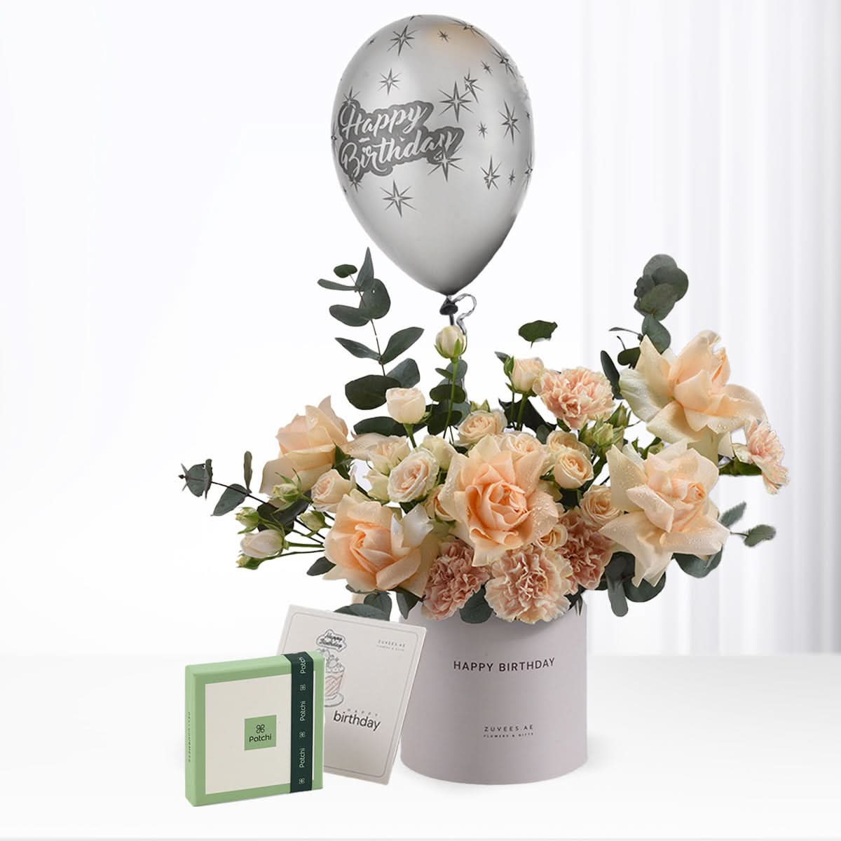 Silver balloon with stars floats above peach roses in gray cylinder, beside green gift box.