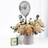 Silver balloon with stars floats above peach roses in gray cylinder, beside green gift box.