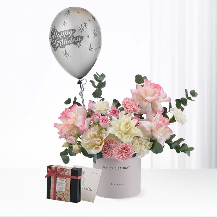 Silver balloon floats above pastel pink/white roses and eucalyptus in a “Happy Birthday” vase, beside a gift box.