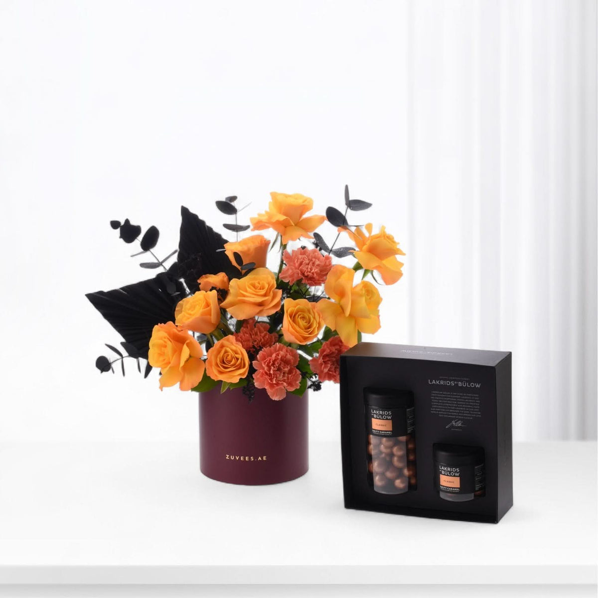 Vibrant orange roses, black foliage in burgundy Zuvees vase beside Lakrids Bülów chocolate gift box.