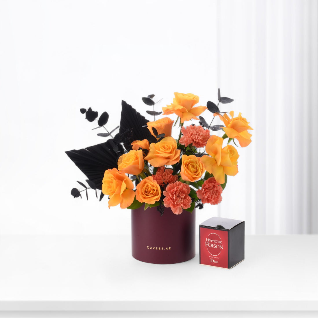 Vibrant orange roses and carnations in burgundy vase beside Dior Hypnotic Poison perfume.