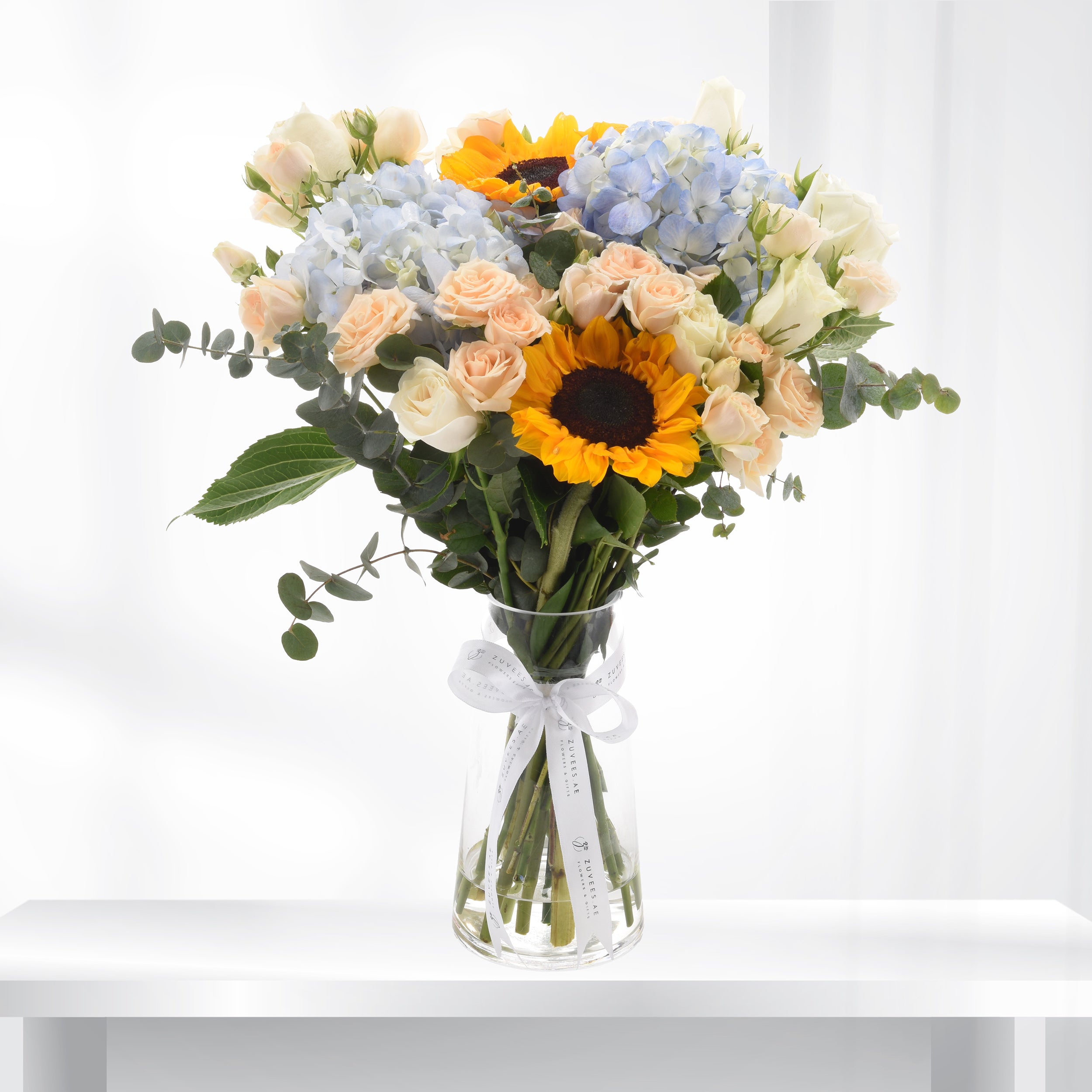 Vibrant sunflowers, blush roses, and blue hydrangeas elegantly arranged in clear glass vase with white ribbon.