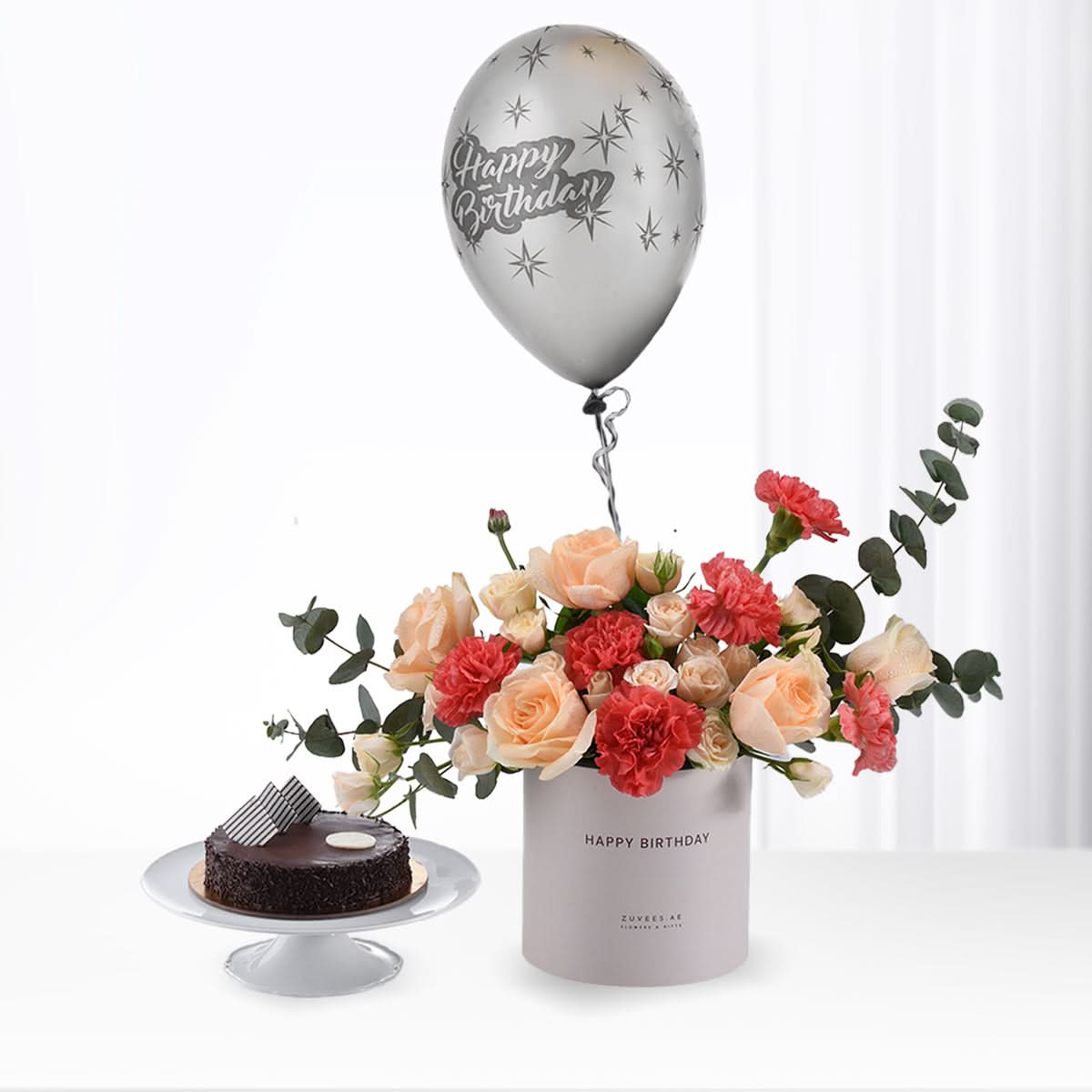 Silver balloon floats above peach and red floral arrangement beside chocolate cake.