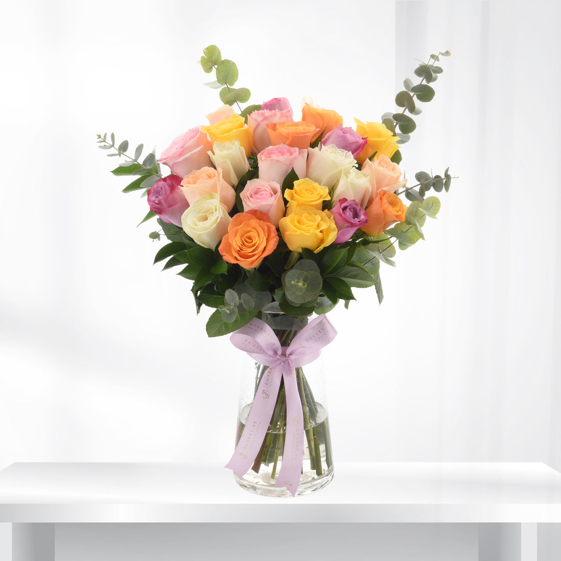 Vibrant multi-colored roses in clear vase, accented with eucalyptus and lavender ribbon.
