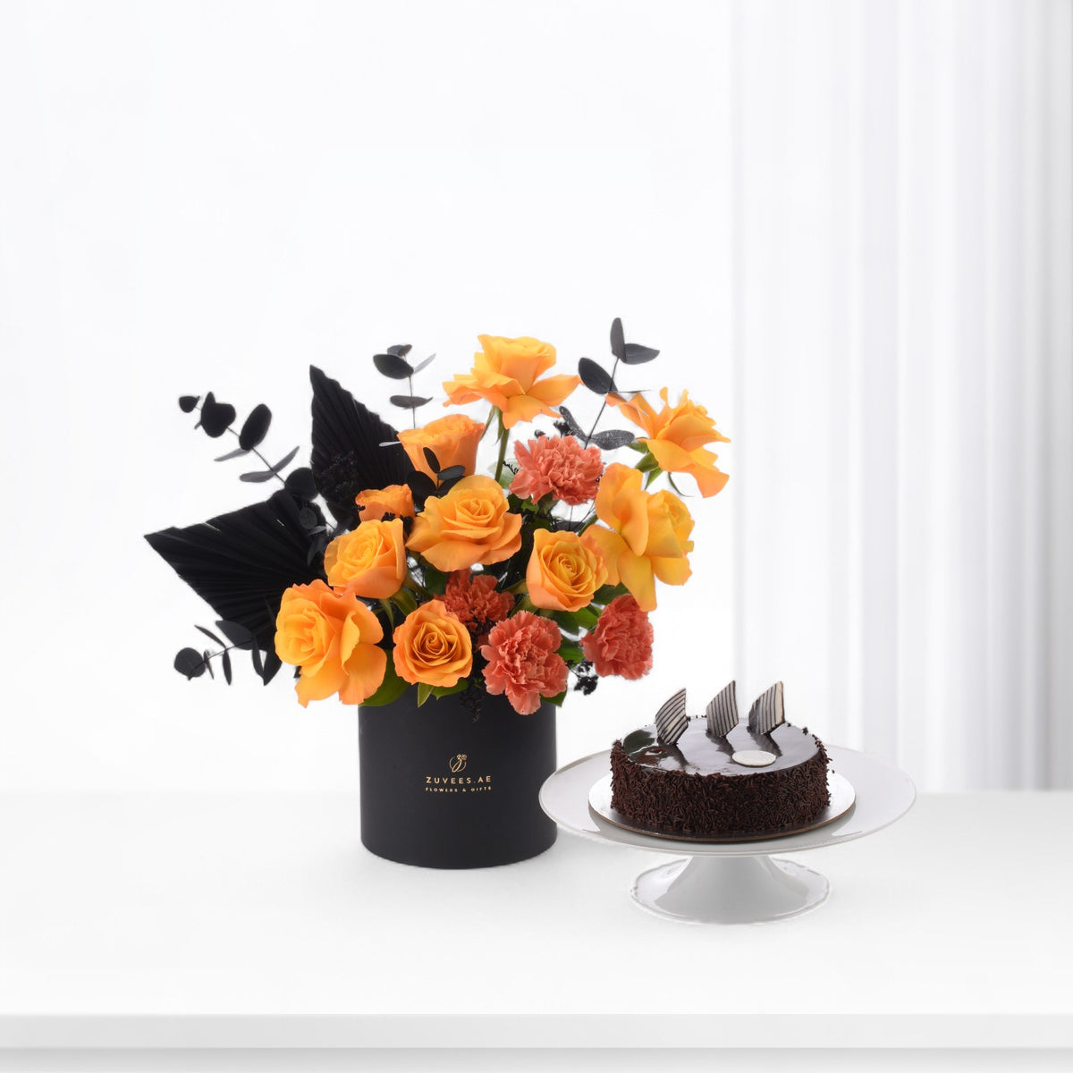 Vibrant orange roses and carnations with black foliage in matte black vase beside chocolate cake.