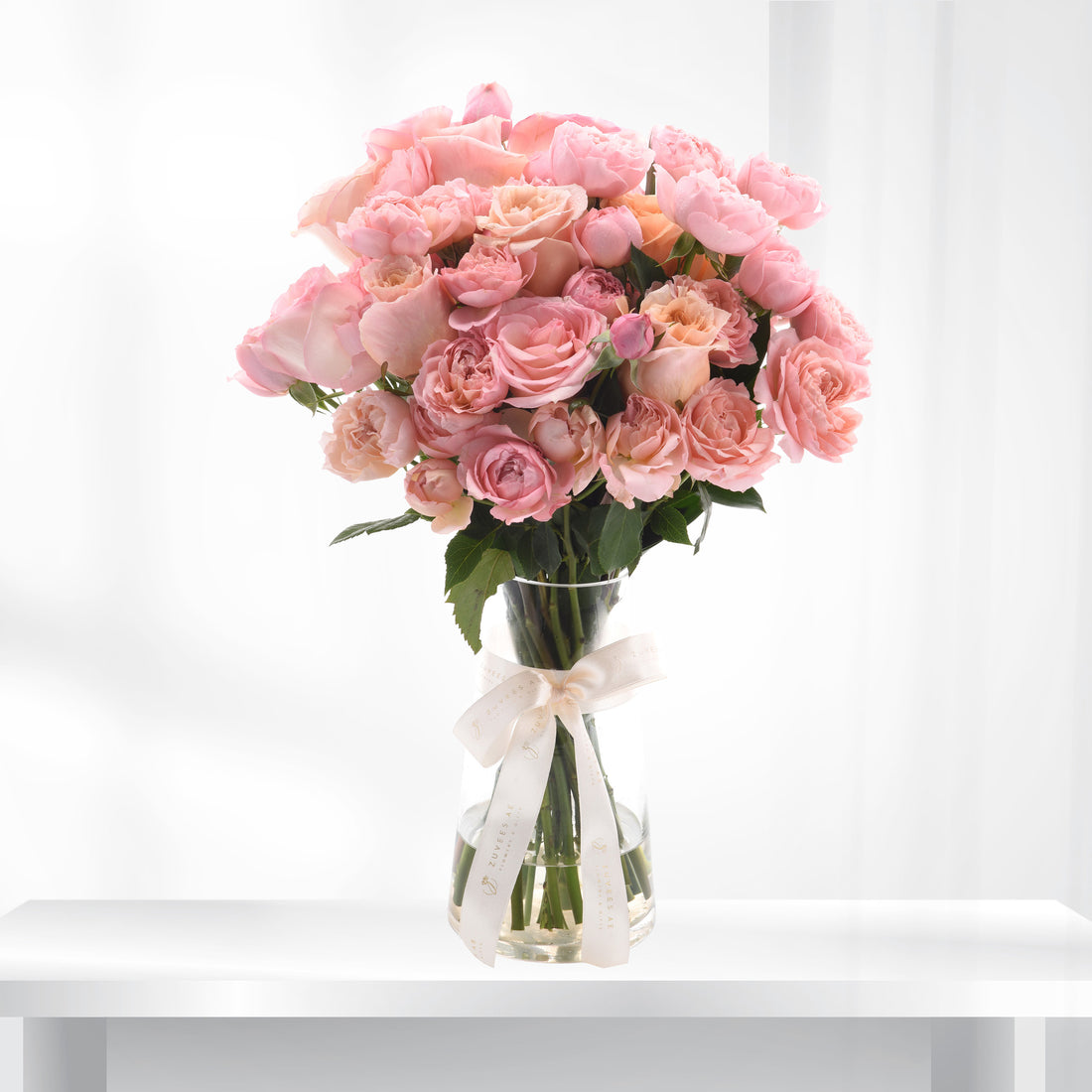 Soft pink roses in clear vase, elegantly tied with cream ribbon.