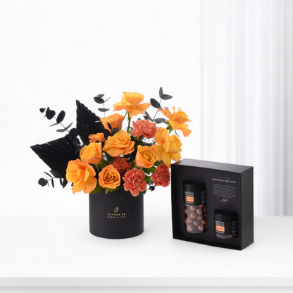 Vibrant orange roses and black foliage in sleek black vase beside gourmet chocolate gift box.