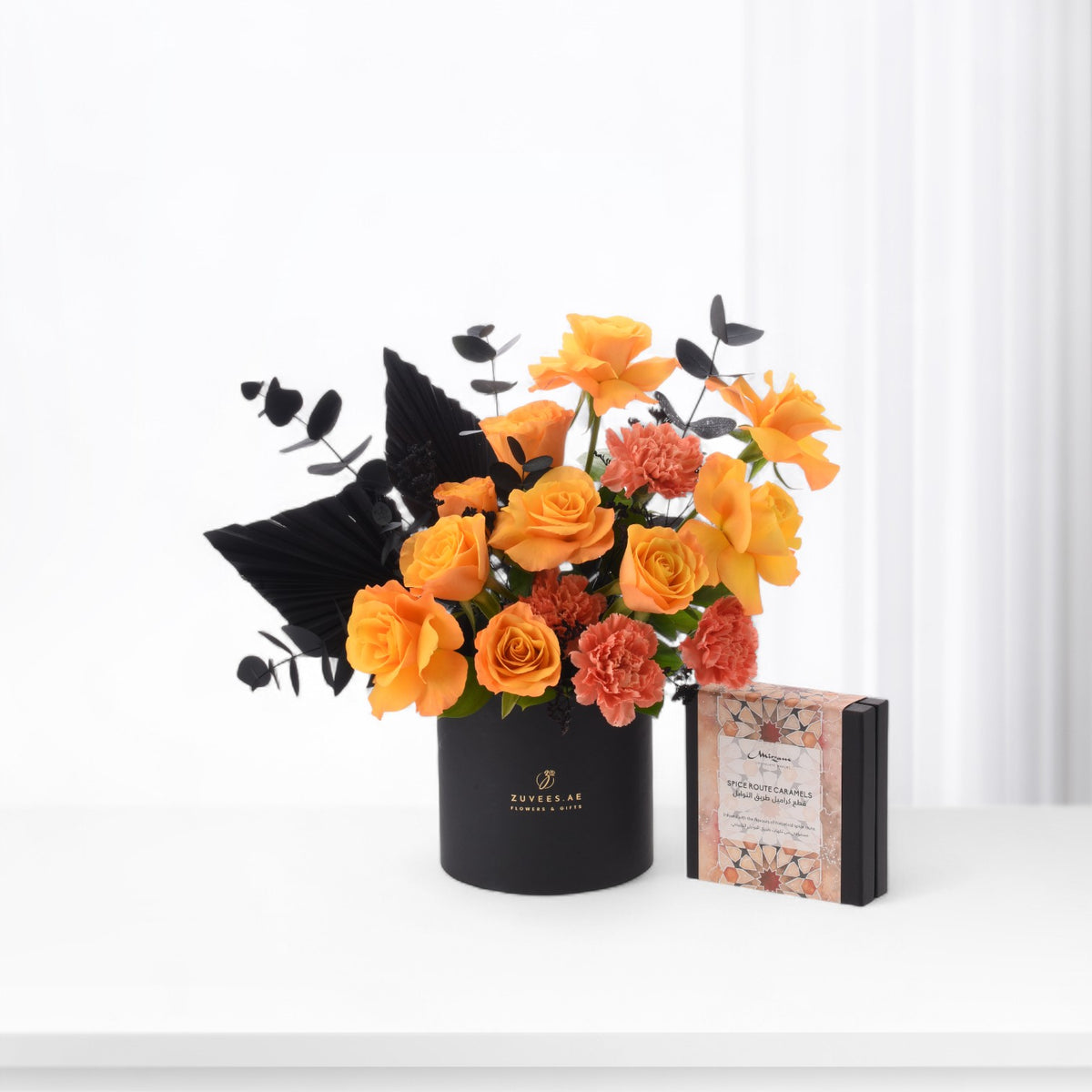 Bold orange roses and coral carnations contrast with black foliage in sleek black vase beside caramel gift box.