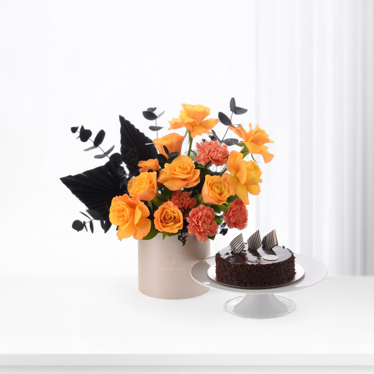 Vibrant orange roses and carnations with black foliage, beside chocolate cake on white stand.