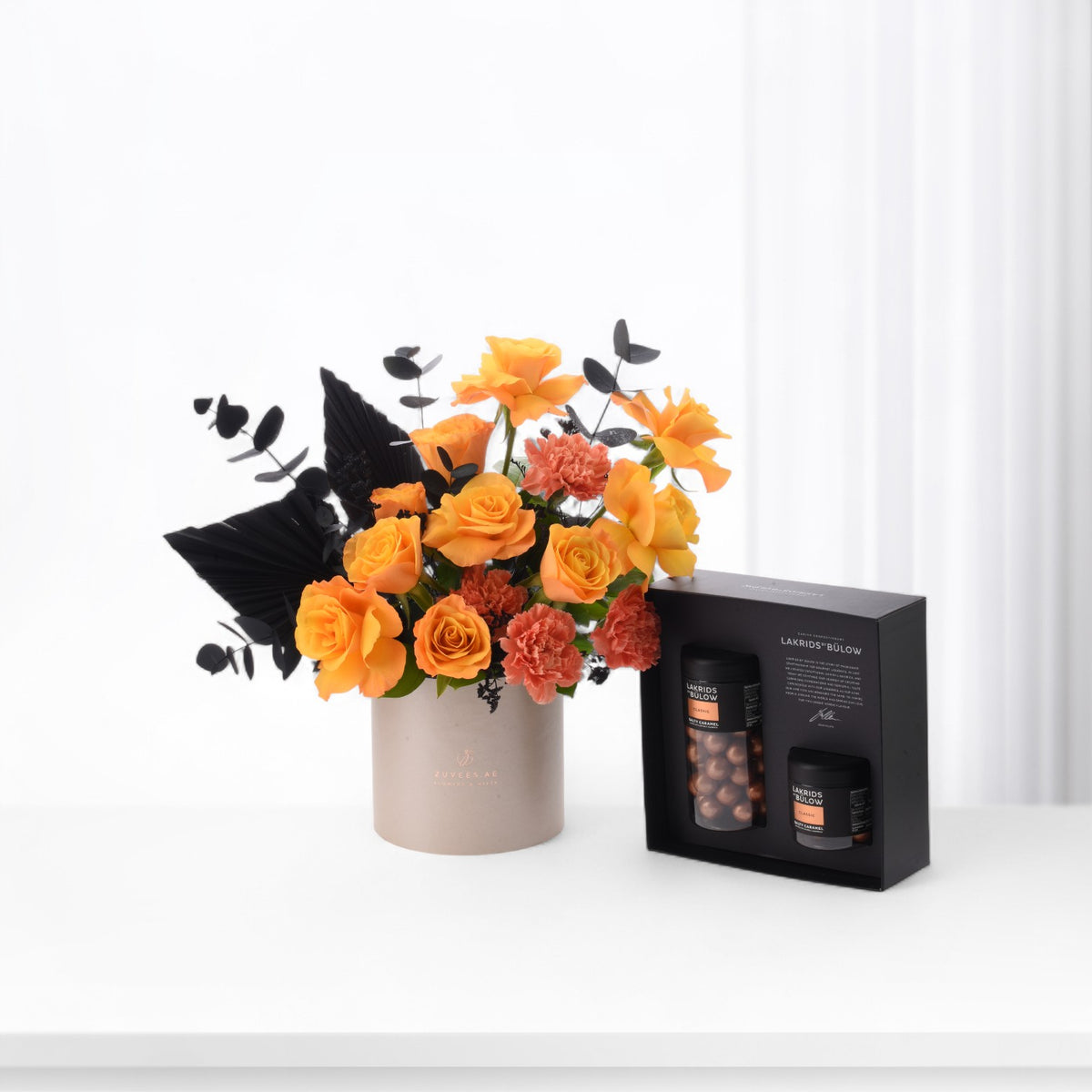 Vibrant orange roses and black foliage in beige vase beside sleek black chocolate gift box.
