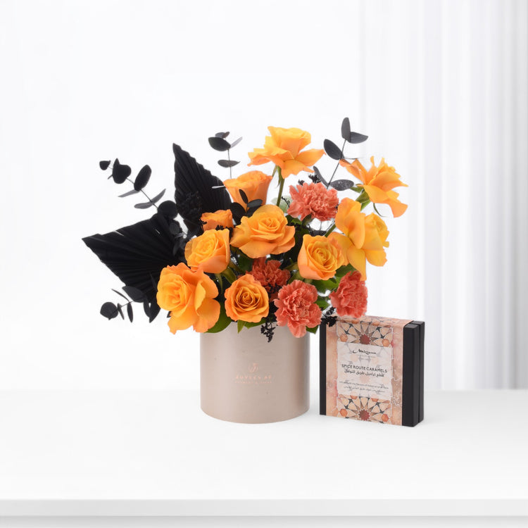 Vibrant orange roses and coral carnations contrasted with dramatic black foliage in minimalist beige vase beside spice caramel gift box.