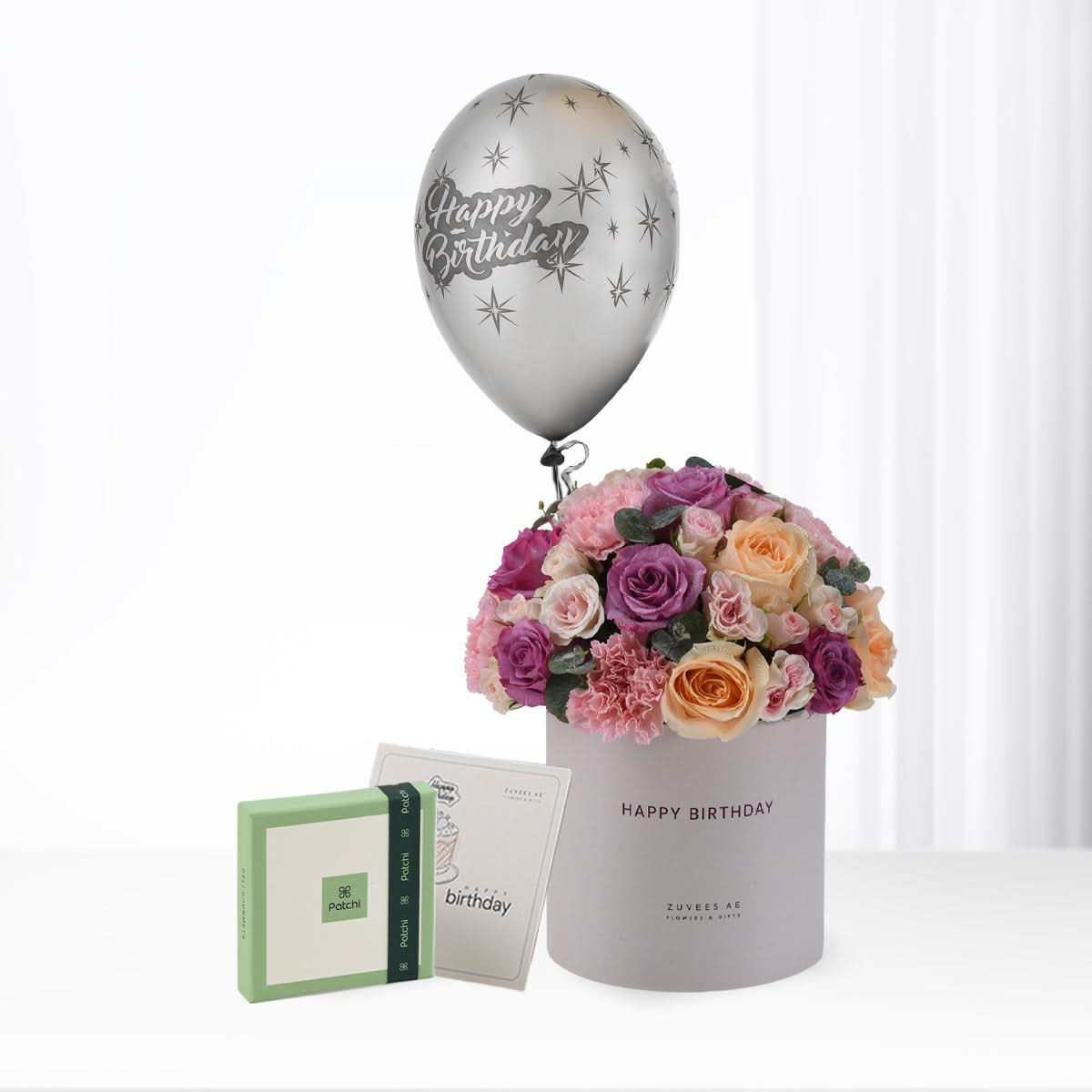 Silver balloon with stars floats above pastel rose bouquet in gray "Happy Birthday" box, beside green Patchi chocolate gift.