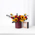 Vibrant red and yellow roses with sunflowers in burgundy vase beside caramel gift box.