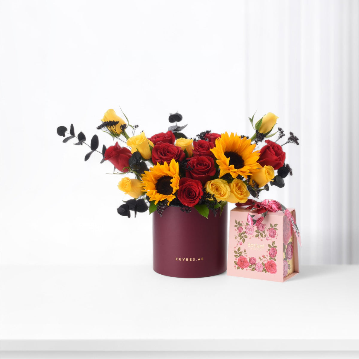 Vibrant red roses, yellow sunflowers, and black eucalyptus in burgundy Zuvees box with pink gift box.