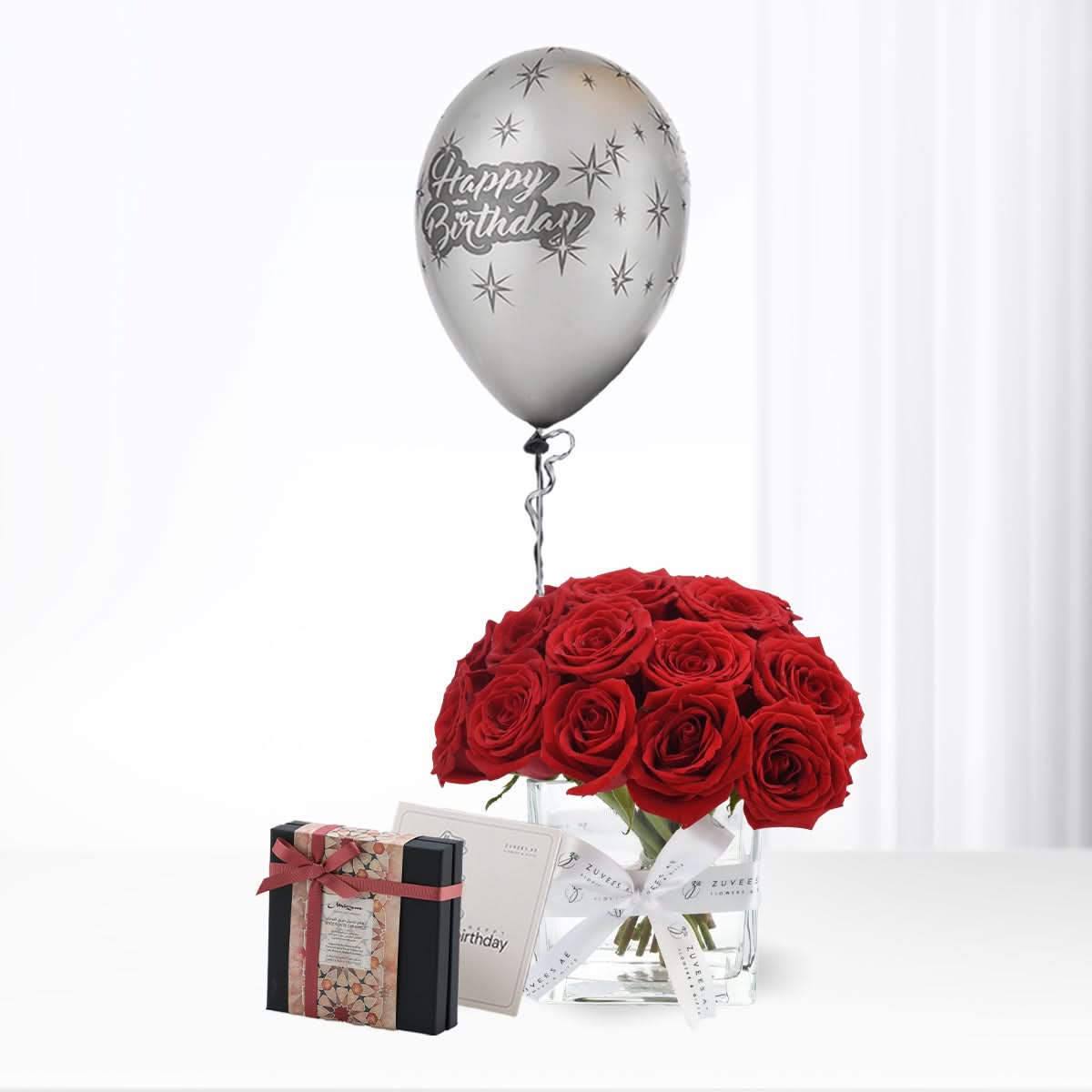 Silver "Happy Birthday" balloon floats above vibrant red roses in clear vase with gift box.
