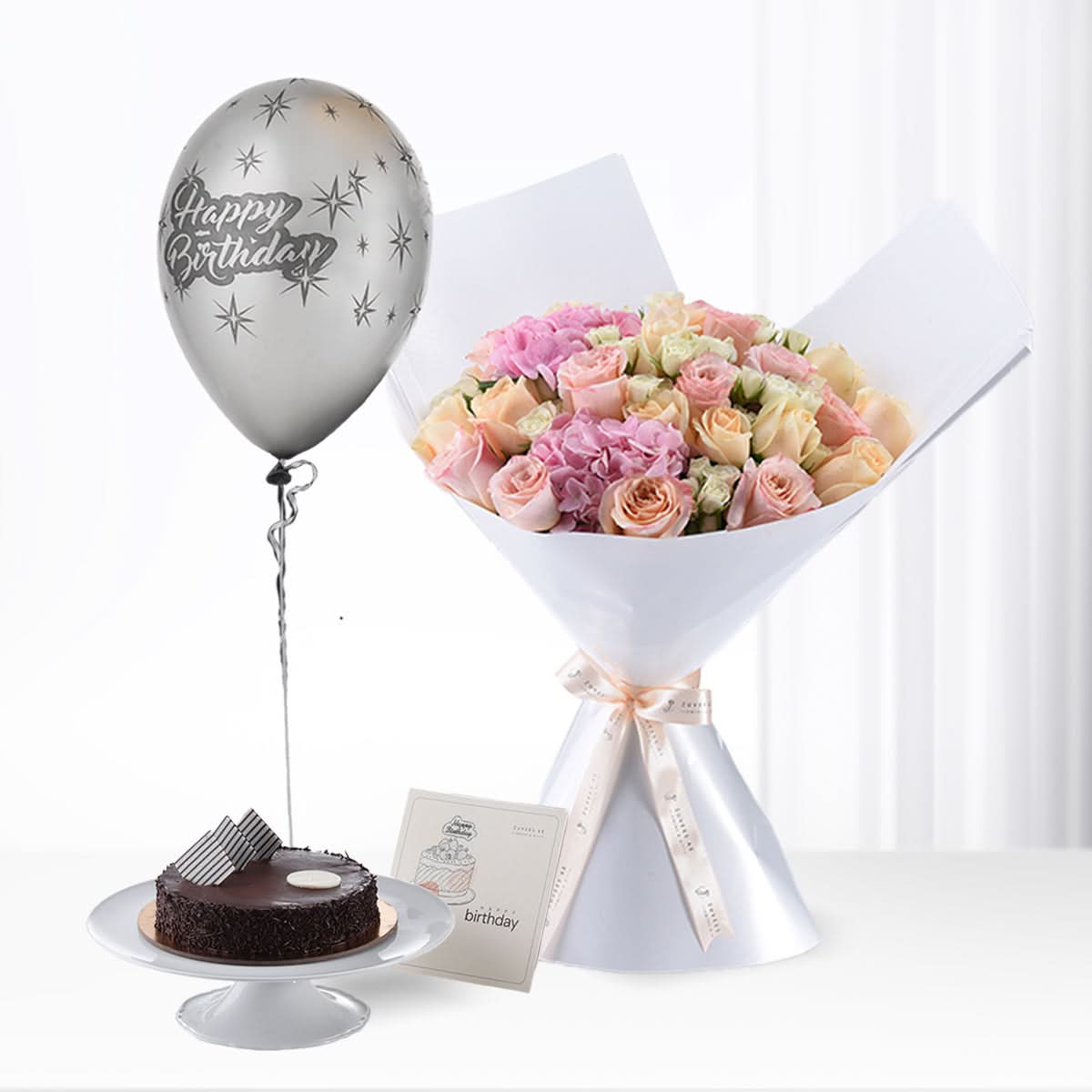 Pastel rose bouquet, silver "Happy Birthday" balloon, chocolate cake, and greeting card on white surface.