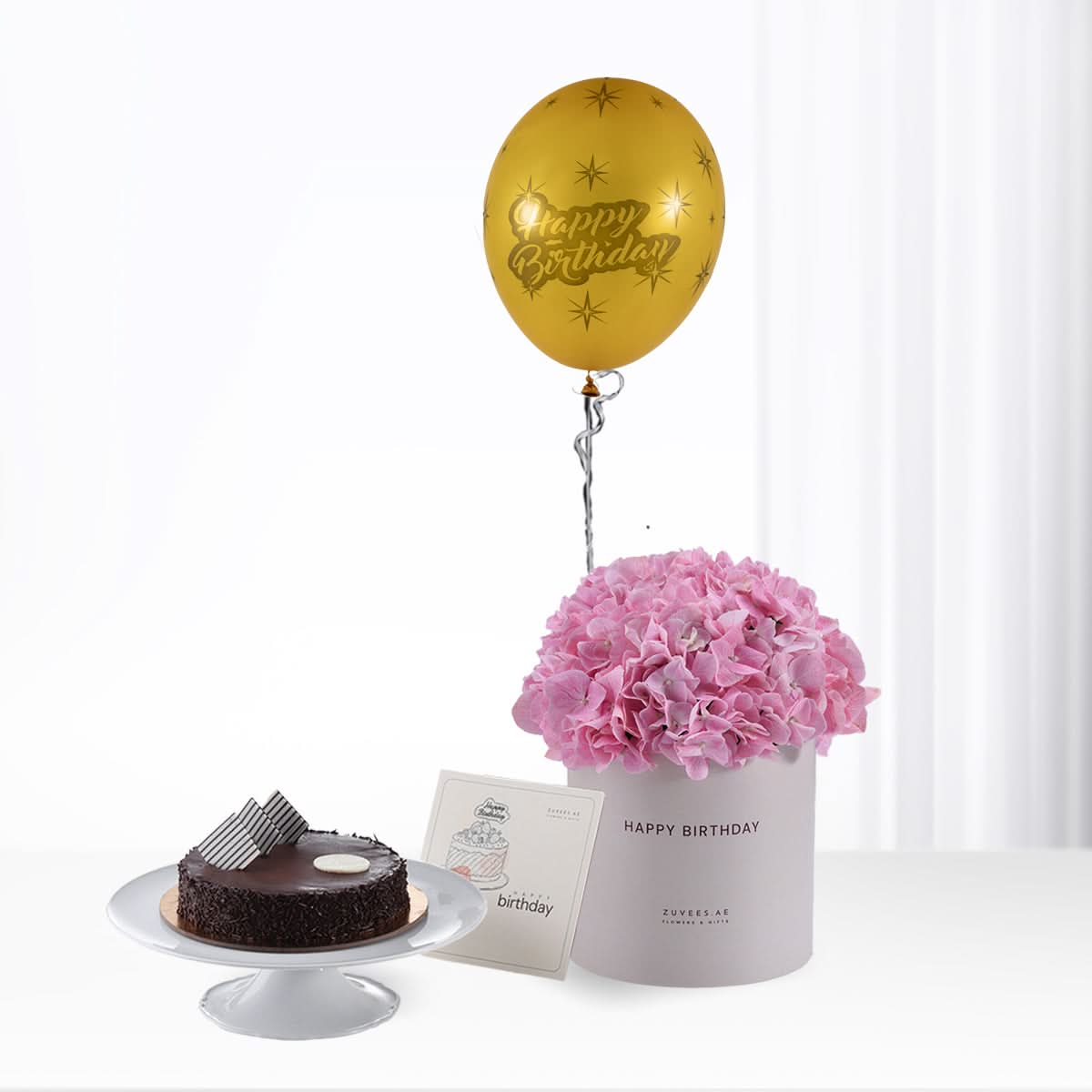 Golden "Happy Birthday" balloon atop pink hydrangeas, beside chocolate cake.