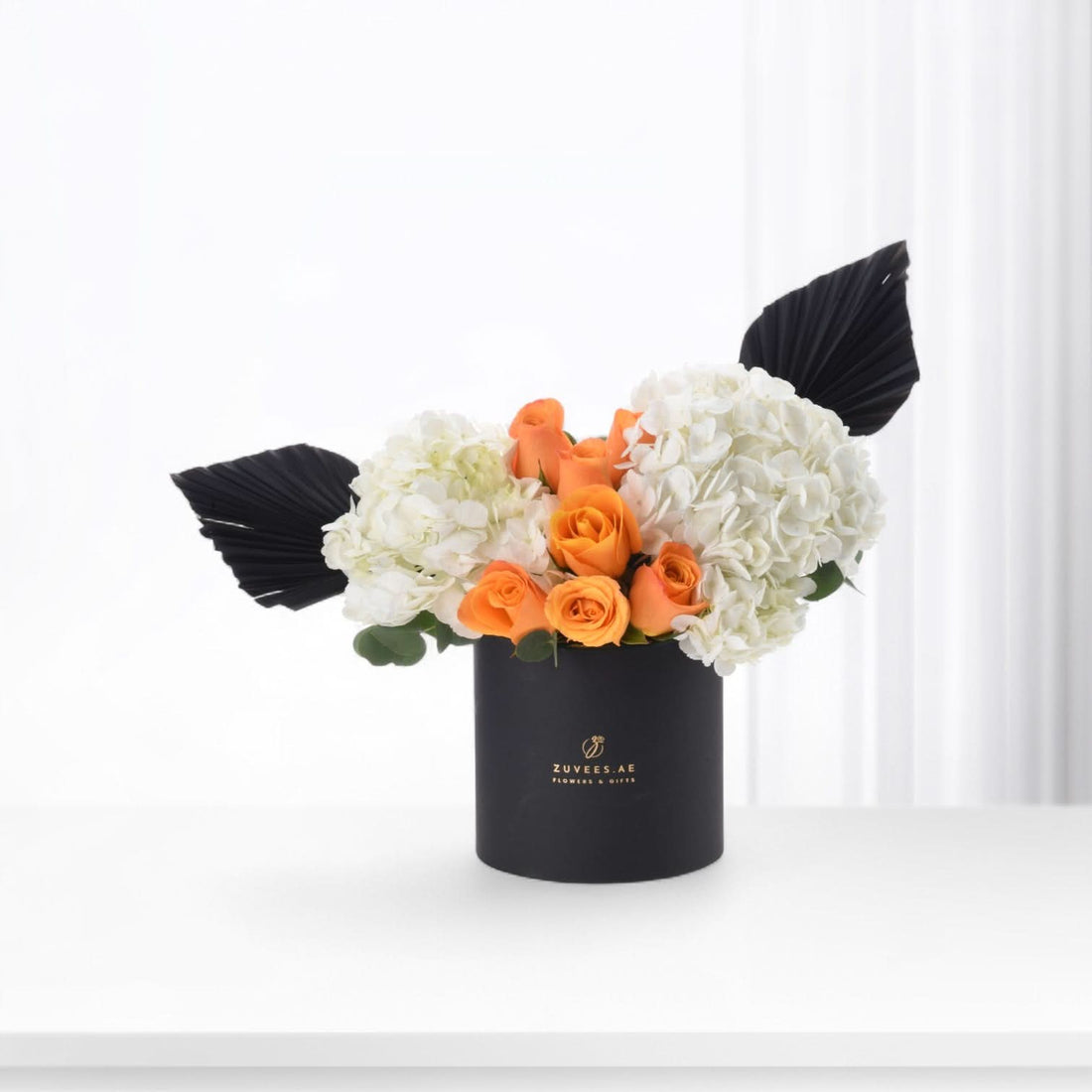 Black cylindrical vase holds vibrant orange roses and white hydrangeas, accented with dramatic black fan leaves.