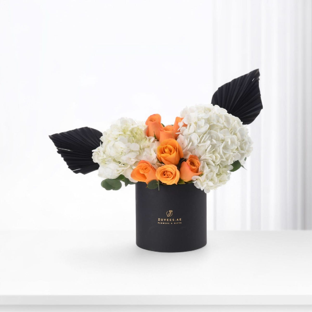 Black cylindrical vase holds vibrant orange roses and white hydrangeas, accented with dramatic black fan leaves.