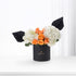 Black cylindrical vase holds vibrant orange roses and white hydrangeas, accented with dramatic black fan leaves.