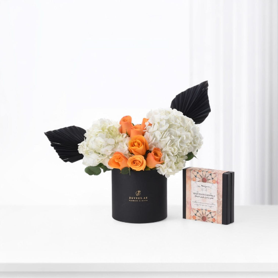 Black vase with orange roses and white hydrangeas, accented by black fan leaves, beside a spice caramel gift box.
