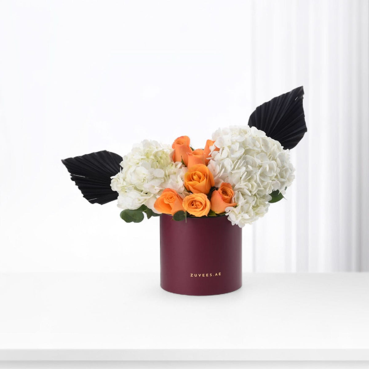 Vibrant orange roses and white hydrangeas in burgundy box with black fan leaves.