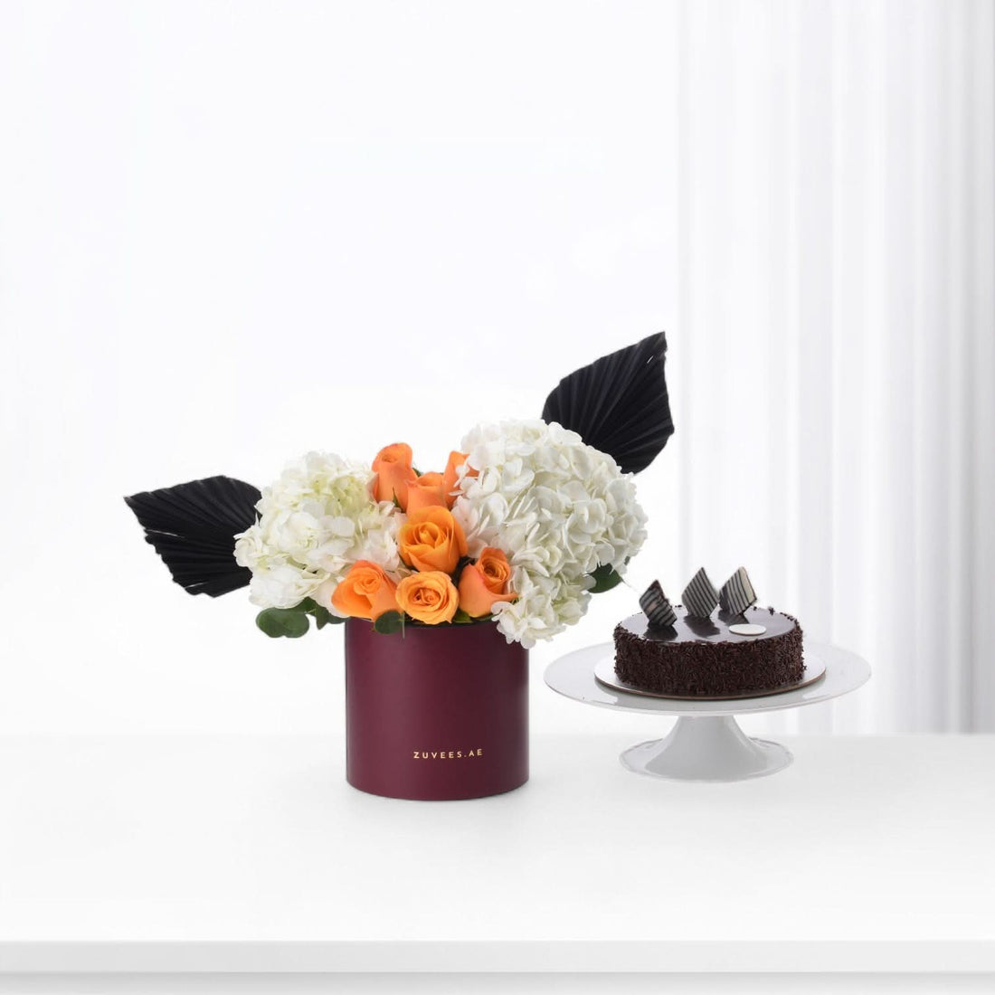 Bold orange roses and white hydrangeas in burgundy box beside chocolate cake.